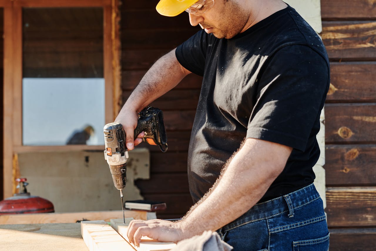 A construction worker wearing a helmet uses a power drill on a wooden plank at an outdoor construction site.