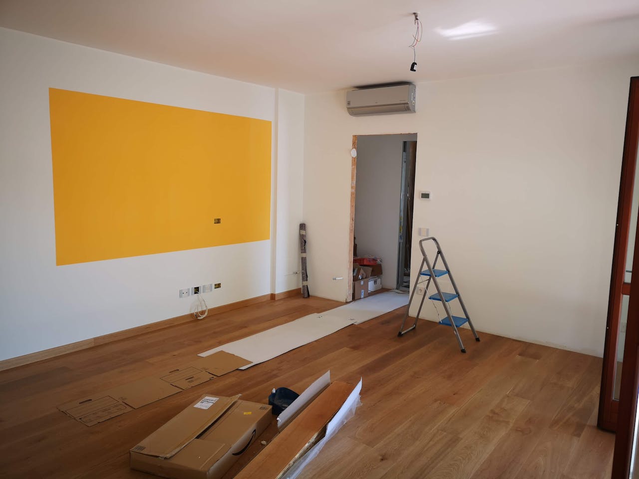 about-03 A bright, empty room under renovation with ladder, tools, and wooden flooring.